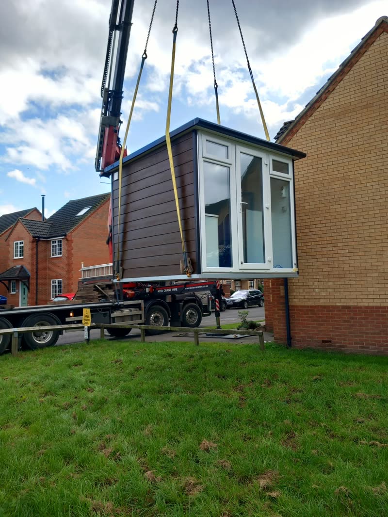 A grey garden room being lifted into place by a lorry crane beside a brick house
